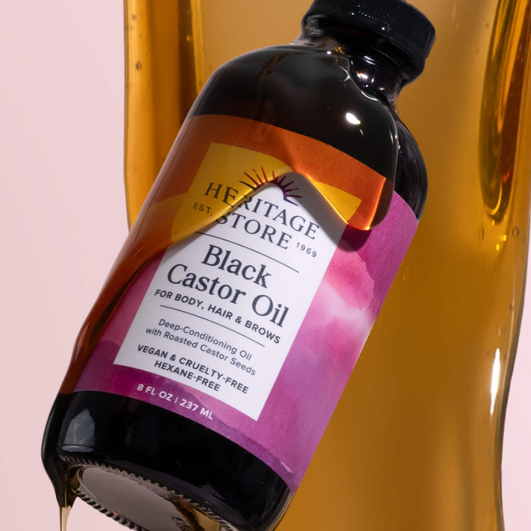A bottle of Heritage Store Black Castor Oil is tilted with oil spilling out. The label mentions its for body, hair, and brows, is vegan, cruelty-free, and hexane-free. Background features flowing oil.